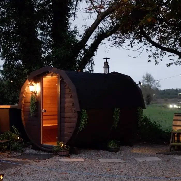 The barrel sauna lit from within at dusk, under the trees in the yard.